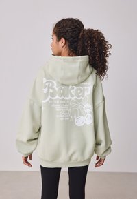 Világoszöld, túméretes kapucnis pulóver puha anyagból. Fehér grafikus szöveg és illusztrációk találhatók a hátoldalán. Bordázott mandzsetták és szegély.