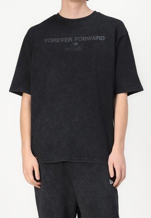 Osoba ubrana w czarny cętkowany t-shirt z napisem "FOREVER FORWARD EST. 1963" w ciemnym nadruku na piersi oraz pasujące czarne spodnie.