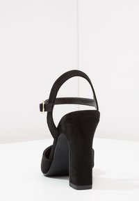 Černé semišové sandály na vysokém podpatku s širokým podpatkem, páskem kolem kotníku a sponou v zlatém odstínu. Hladký povrch s otevřeným zadním designem.