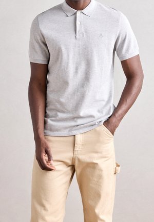 Homme portant un polo gris clair à manches courtes et un pantalon beige, une main dans la poche, debout devant un fond clair uni.