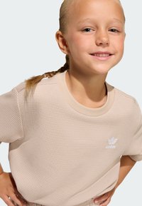 Béžový pletený crop top s textúrou, okrúhlym výstrihom a krátkymi rukávmi. Na ľavej strane na hrudi je malá biela značka Adidas.