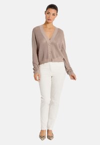 Hellbrauner, gestrickter Cardigan mit tiefem V-Ausschnitt und drei Knöpfen, kombiniert mit weißen Hosen und gemusterten Schuhen. Strukturiertes Gewebe.