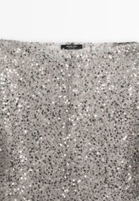 Tessuto di paillettes argentate con una superficie texturizzata, caratterizzato da piccole paillettes rotonde in argento e nero, che creano un motivo riflettente. Etichetta del marchio visibile.