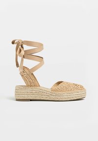 Sandale compensée espadrille avec dessus en raphia tissé, rubans beige à nouer autour de la cheville et semelle plateforme en jute tressée.