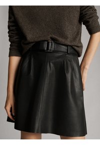 Sort lædernederdel med folder, der har et bredt bælte. Nederdelen har en glat overflade og en svungen form, og den bæres med en brun sweater.