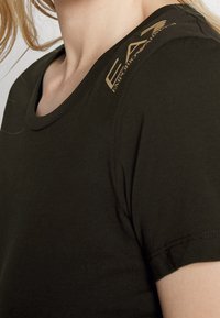 Tmavě zelené tričko s kulatým výstřihem, krátkými rukávy a potiskem zlatého loga na rameni. Měkký bavlněný materiál s hladkým povrchem.