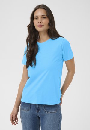 Mujer con cabello largo y oscuro, vistiendo una camiseta de manga corta azul brillante y jeans azules, de pie frente a un fondo claro y liso.