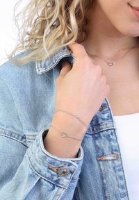 Silberne Halskette mit Herzanhänger und Silberne Kettenarmband. Getragen über einem weißen Oberteil und einer Jeansjacke, die ein filigranes Design zeigt.