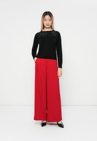 Sort langærmet top med pailletdetaljer ved halsen, parret med brede røde bukser. Sorte spidse sko fuldender outfittet.
