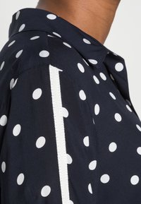 Camicia blu navy con pois bianchi, dotata di colletto e di una striscia verticale bianca strutturata lungo la cucitura della spalla.