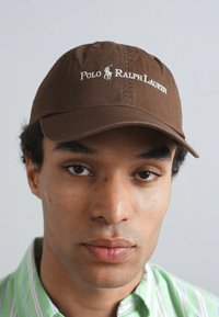 Rjav bombažni baseball klobuk z belim logotipom "Polo Ralph Lauren", klasično ukrivljenim vizirjem in nastavljivim hrbtnim trakom.