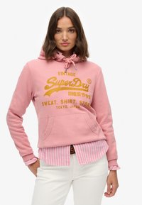 Roze hoodie gemaakt van zachte stof met een voorzak, voorzien van een gouden logo en tekst. Gedragen over een roze en witte gestreepte shirt.