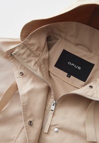 Giacca impermeabile beige con chiusura a zip, caratterizzata da una texture liscia e un'etichetta interna contrassegnata "OPUS". Include bottoni a scatto e un cappuccio.