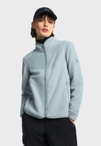 Svijetloplava flis jakna s visokim ovratnikom, punim zatvaračem i bočnim džepovima. Sadrži kontrastne detalje i logo brenda na rukavu.