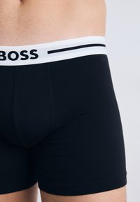 Čierne boxerky vyrobené z mäkkej tkaniny, s bielym elastickým pásom s logom "BOSS" a dvomi čiernymi horizontálnymi pruhmi.