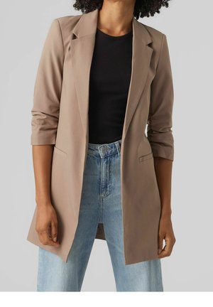 Frau trägt einen beigen Blazer mit hochgekrempelten Ärmeln, schwarzes geripptes Shirt und hellblaue hoch taillierte Jeans vor einem schlichten Hintergrund.