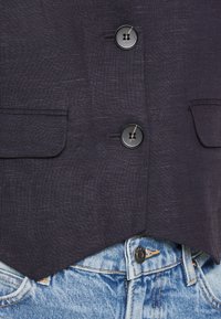 Marineblaues Leinenwest mit zwei schwarzen Knöpfen und aufgesetzten Taschen, kombiniert mit hellblauen Jeans aus Denim mit Metallknopf und kontrastierenden Nähten.