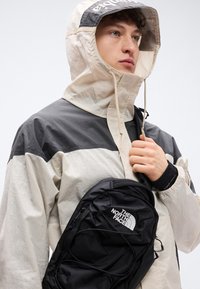 Μαύρο σακίδιο πλάτης από νάιλον με το λογότυπο The North Face, φορεμένο στο στήθος πάνω από ένα μπουφάν με μπεζ και γκρι χρωματικούς τομείς και κουκούλα.