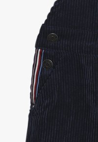 Tmavě modrá kordurová látka s vertikálním žebrováním, zaobleným tvarem a černými knoflíky s vícebarevným pruhovaným akcentem.