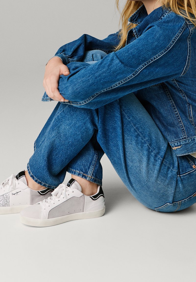 Personne assise en tailleur portant une veste en jean bleu et un jean avec des baskets blanches et grises à lacets sur un fond neutre.