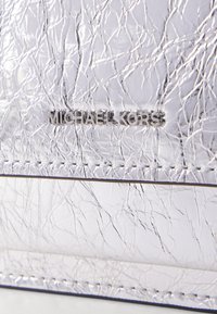 Stříbrný strukturovaný syntetický materiál s nařaseným povrchem. Obsahuje vyražené logo "MICHAEL KORS" a viditelné švy.