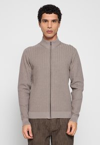 Vest met ritssluiting in lichtbruine gebreide stof, met geribbelde textuur, hoge kraag en lange mouwen. Details omvatten een ritssluiting aan de voorkant en aansluitende boorden.