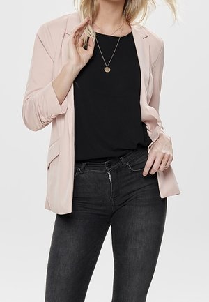 Vrouw die een lichtroze blazer draagt over een zwart topje, gecombineerd met hooggerande donkergrijze spijkerbroek, geaccentueerd met een ronde hangerketting.