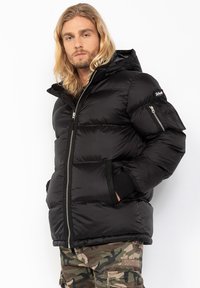 Sort pufferjakke med lynlås foran, hætte og elastiske manchetter. Har en horisontal quiltet design og en sidelomme med lynlås.