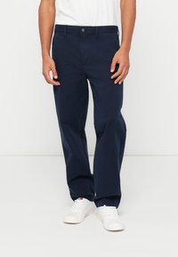 Ναυτικά μπλε βαμβακερό παντελόνι με χαλαρή εφαρμογή, που διαθέτει κούμπωμα με κουμπί, πλάγιες τσέπες και απλή στρίφωση. Συνδυασμένο με λευκά αθλητικά παπούτσια.