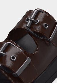 Κοντινό πλάνο σκούρου καφέ δερμάτινου σανδαλιού με δύο ρυθμιζόμενες ασημένιες πόρπες και παχιά μαύρη σόλα.