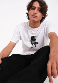 T-shirt en coton blanc avec un graphisme noir d'un oiseau en position debout, associé à un pantalon noir ample. Collier en argent inclus.