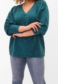 Sweter dziergany w kolorze morskim, z dekoltem w serek, długimi bufkami i prążkowanym obszyciem, zestawiony z dopasowanymi jasnoszarymi jeansami. Miękka faktura i luźny krój.