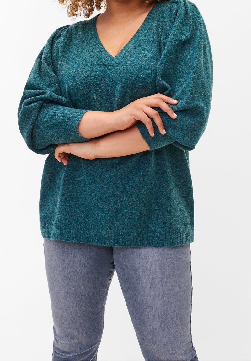 Sweter dziergany w kolorze morskim, z dekoltem w serek, długimi bufkami i prążkowanym obszyciem, zestawiony z dopasowanymi jasnoszarymi jeansami. Miękka faktura i luźny krój.