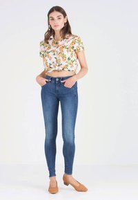 Blommig kortärmad skjorta med orange mönster, i kombination med tajta mörkblå jeans och bruna vävda skor, vilket visar en avslappnad stil.