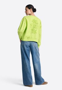 Neonowo zielona bluza o luźnym kroju, z nadrukiem "SUPER WOMAN" i koniczynkami na plecach, zestawiona z szerokimi dżinsami o prostych nogawkach.
