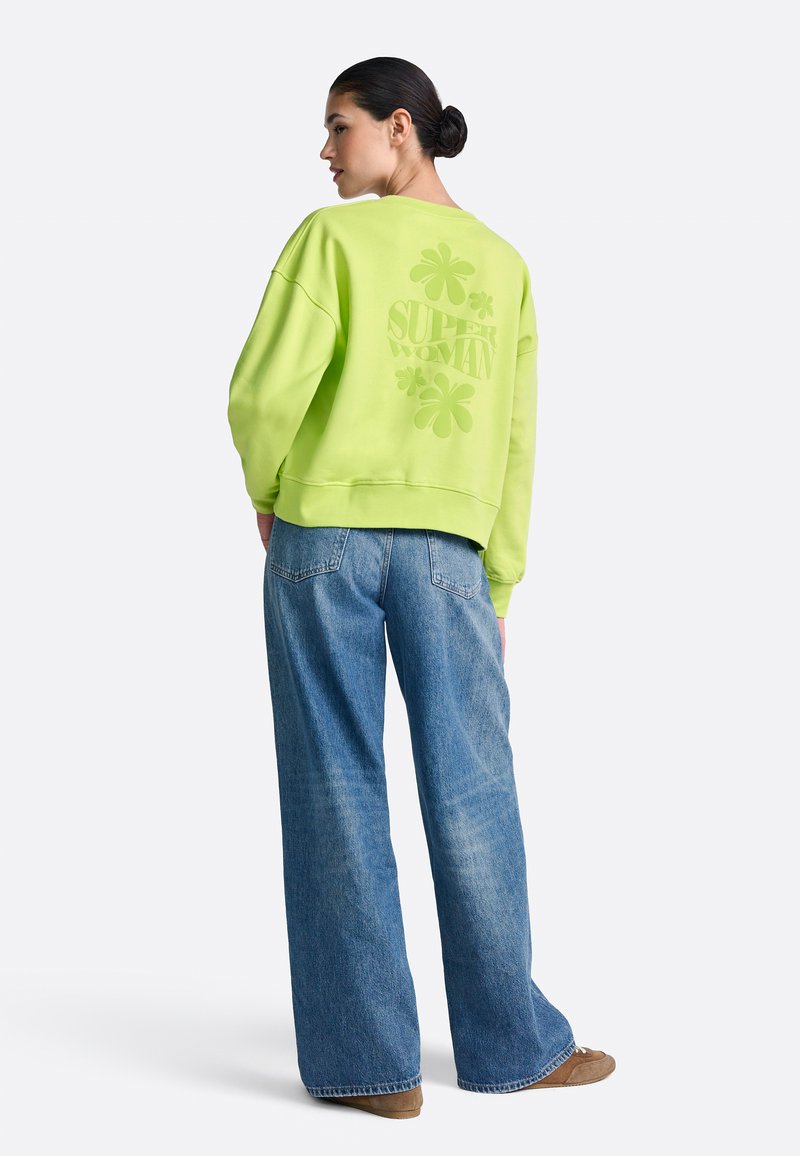 Neonowo zielona bluza o luźnym kroju, z nadrukiem "SUPER WOMAN" i koniczynkami na plecach, zestawiona z szerokimi dżinsami o prostych nogawkach.