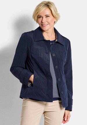 Vrouw draagt een marineblauw jasje met knopen over een marineblauw-wit gestreept shirt, beige broek, glimlacht met één hand in haar jaszak.