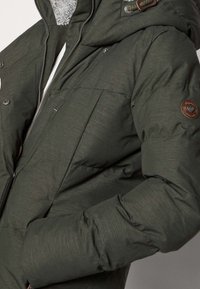 Přetlaková bunda v tmavě zelené tkanině, s uzavíráním na zip, cvoky, prošívaným designem a kapucí podšitou fleecem s logovou nášivkou.