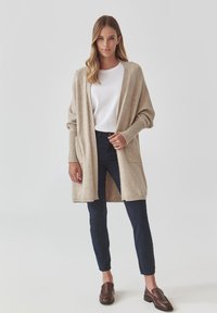 Cardigan en tricot beige avec un devant ouvert, des poignets côtelés et deux poches avant, porté sur un haut blanc et un pantalon foncé. Texture lisse et coupe décontractée.
