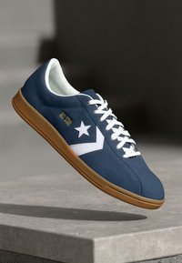 Zapatilla azul marino con cordones blancos, logo de estrella y chevrón blancos, suela marrón y texto "All Star" flotando sobre una superficie de concreto.
