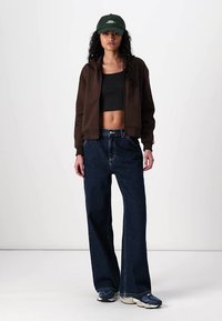 Brun zip-up hoodie över en svart crop top, kombinerad med lösa mörkblå jeans och marinblå sneakers. En grön keps fulländar outfiten.