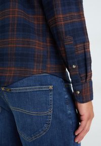 Geruite flanellen shirt in marineblauw en gebrande oranje, met knoopmanchetten en een afgeronde zoom, gecombineerd met blauwe spijkerbroek met gele stiksels.