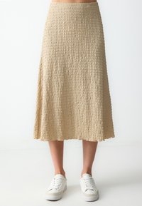 Beige, knielanger Rock aus strukturiertem Stoff mit gewelltem Saum. Kombiniert mit weißen Sneakers.