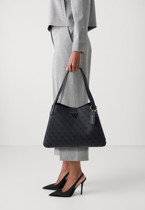 Personne portant un costume à carreaux noir et blanc et des escarpins noirs, tenant un grand sac à main Guess noir avec logo et étiquette.