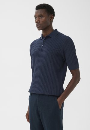 Mann trägt ein marineblaues, kurzärmeliges, kabelgestricktes Poloshirt und dazu passende Hosen, steht mit einer Hand in der Tasche vor schlichtem Hintergrund.