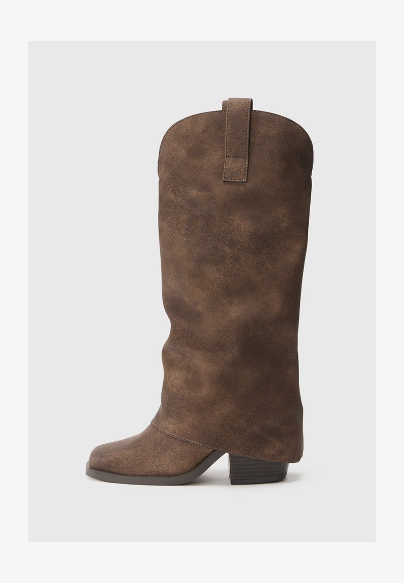 Bota castanha de imitação de pele até ao joelho com ponta redonda, salto baixo e uma alça na parte de trás. Textura suave com variações subtis.