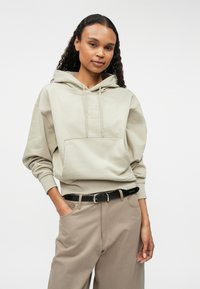 Μπεζ φούτερ με κουκούλα, τσέπη μπροστά, κορδόνι στην κουκούλα και ανάγλυφο λογότυπο. Συνδυασμένο με ανοιχτό καφέ παντελόνια και μαύρη ζώνη. Λείος υφή.