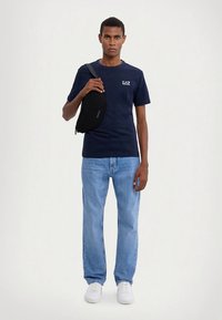 Tmavě modré tričko s krátkým rukávem s logem "EAZ", světlé modré džíny, bílé tenisky a černá kabelka přes rameno, umístěné na neutrálním pozadí.