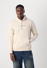 Hoodie color crema con coulisse e tasca anteriore, con il logo "Marc O'Polo DENIM" in testo sottile, abbinato a jeans blu.