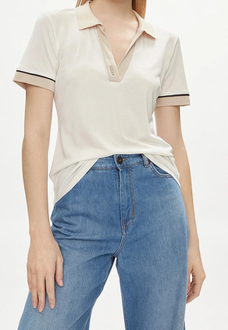 Femme portant un polo crème avec des bordures beige et noire, associé à un jean en denim bleu taille haute sur fond blanc.